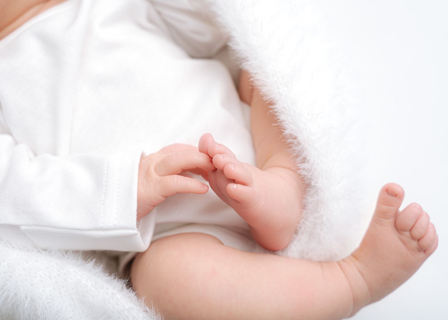A newborn baby's tiny hand reaches out and grasps a finger against a soft white background.