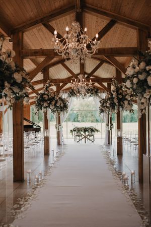 Elegant wedding aisle in rustic wooden venue with crystal chandeliers and white floral arrangements leading to outdoor view.