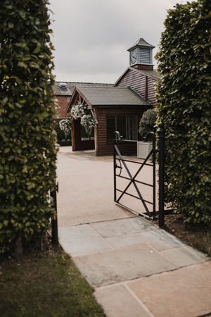 Rustic wooden chapel entrance with black metal gate framed by manicured hedges under overcast sky.