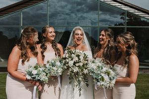 Bridesmaids in blush pink dresses holding white and green bouquets stand together at an outdoor wedding venue.