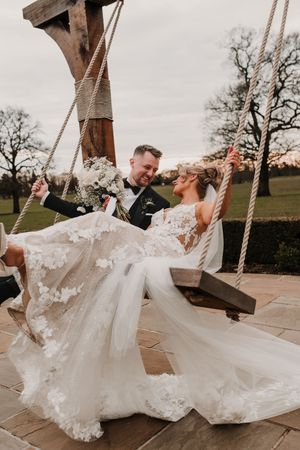 Newlyweds share a joyful moment together on a rustic wooden swing during sunset at their outdoor wedding celebration.