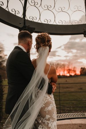 Newlywed couple embraces during sunset at rustic outdoor wedding venue with ornate iron railings at Merrydale Manor