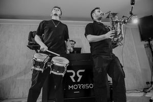 Black and white photo of musicians performing with drums and bagpipes on stage with RS Morriso banner.