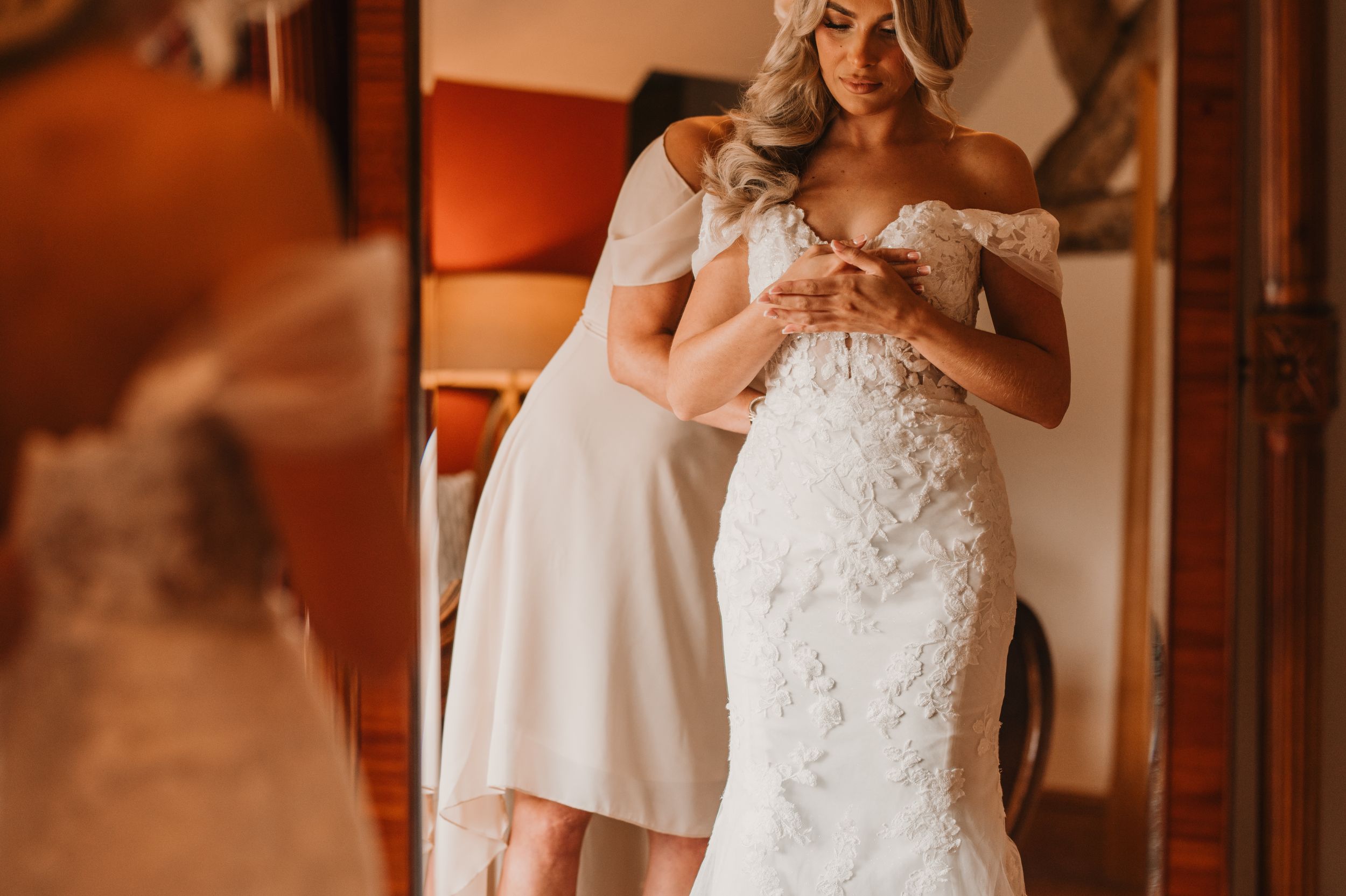 A bride in an ornate lace wedding gown gets help with final adjustments in a warm-lit dressing room at Merrydale Manor