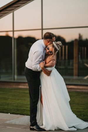 Romantic wedding couple embraces during sunset at outdoor venue with glass building and green lawn.