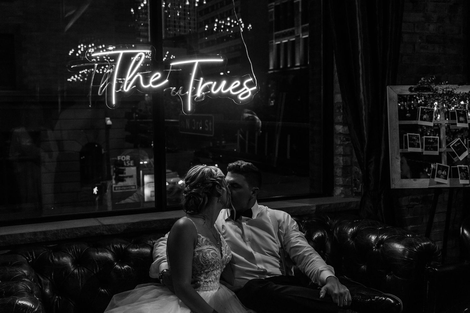 A romantic couple sits together beneath a glowing neon sign reading 'The Tries' in a moody black and white photograph.