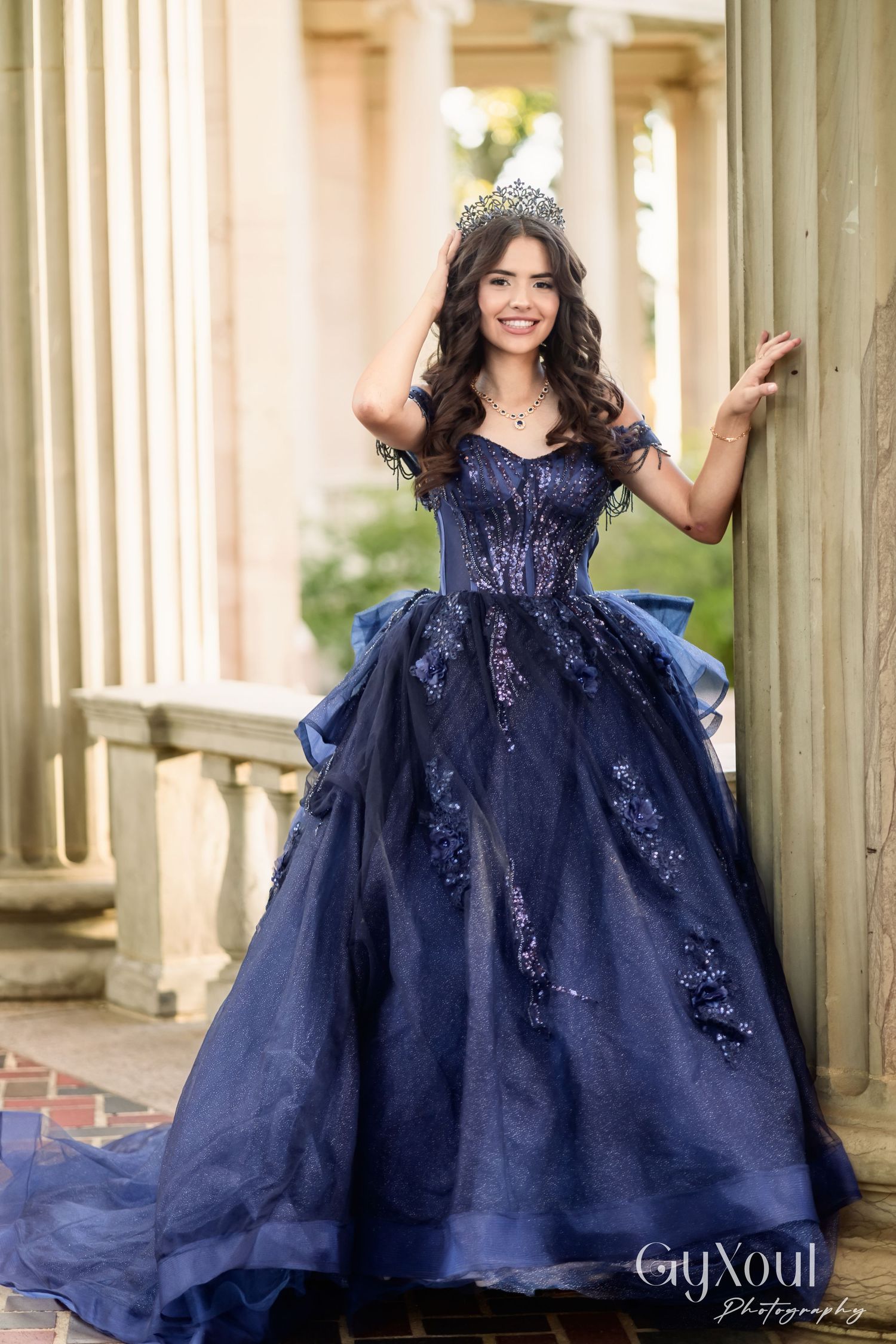 Young woman in a navy blue ball gown with sequins poses in front of wooden pillars in an outdoor setting.