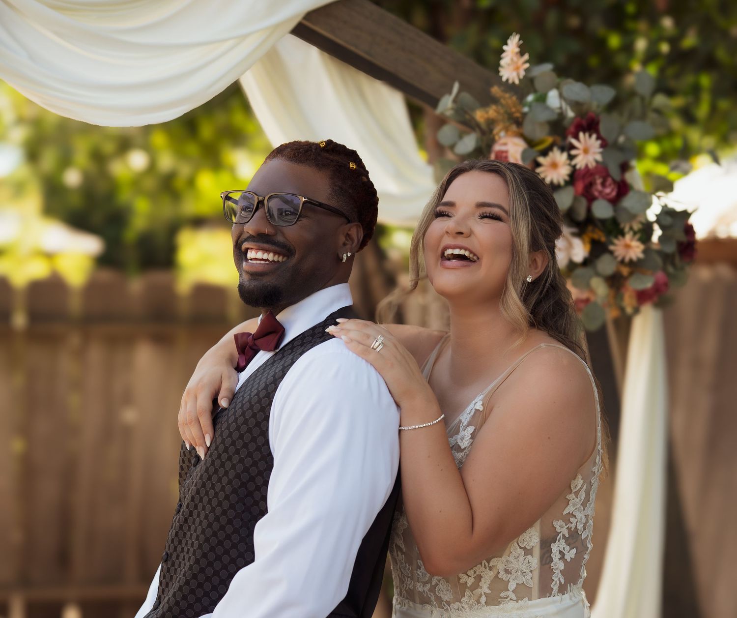 A joyful bride and groom share a candid laughing moment together under a draped wedding ceremony arch with floral decor.