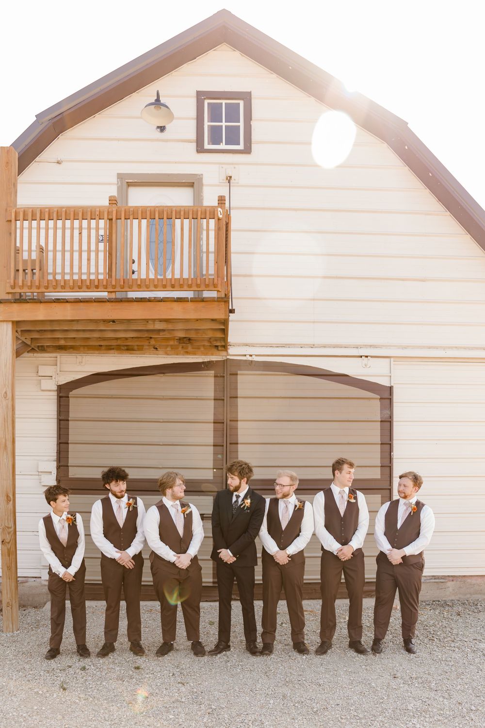 A row of groomsmen in brown vests lean against a white barn wall in matching attire.
