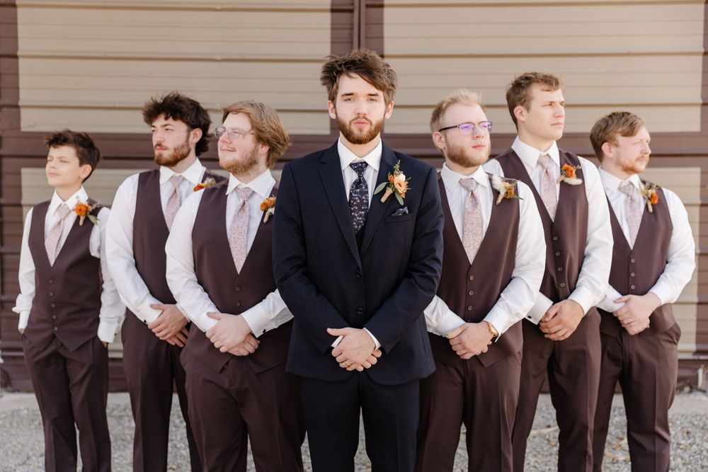 Wedding party members in brown suits and formal attire pose together in a line against a beige wall.