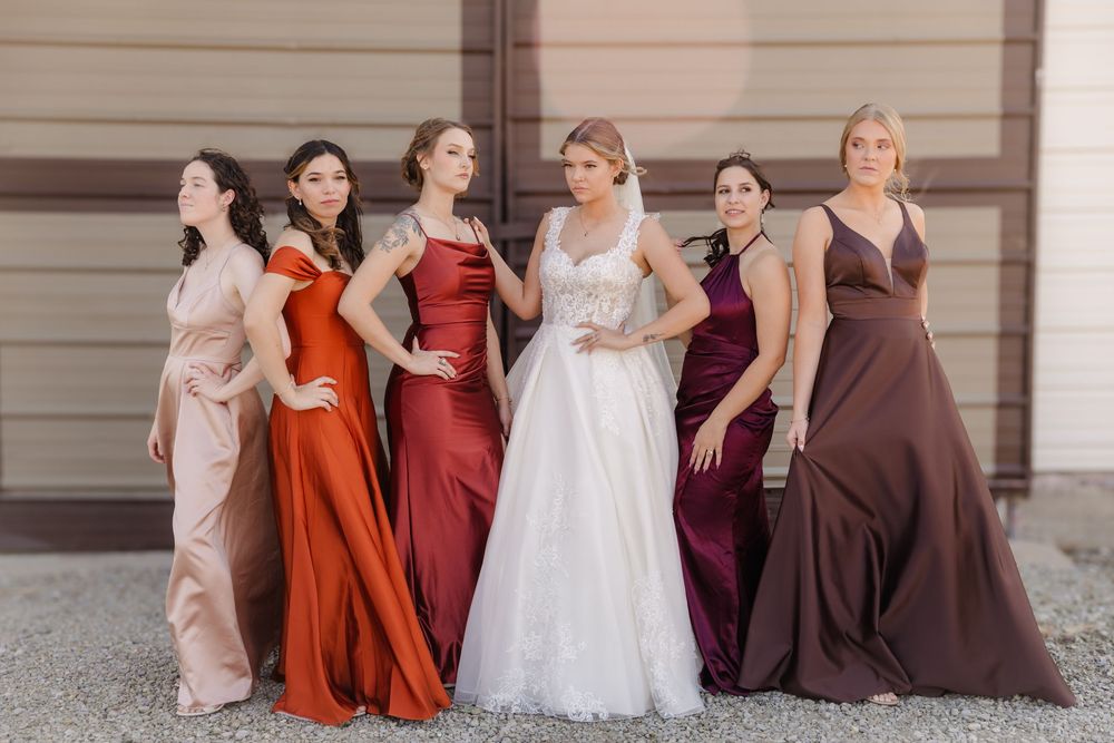 Bridesmaids in burgundy and rust-colored dresses stand with bride in white gown against neutral building exterior.