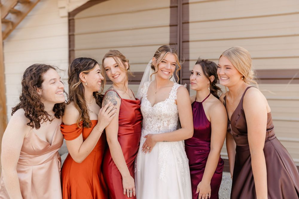 A group of bridesmaids in colorful dresses stand in a line against a beige wall during a wedding celebration.