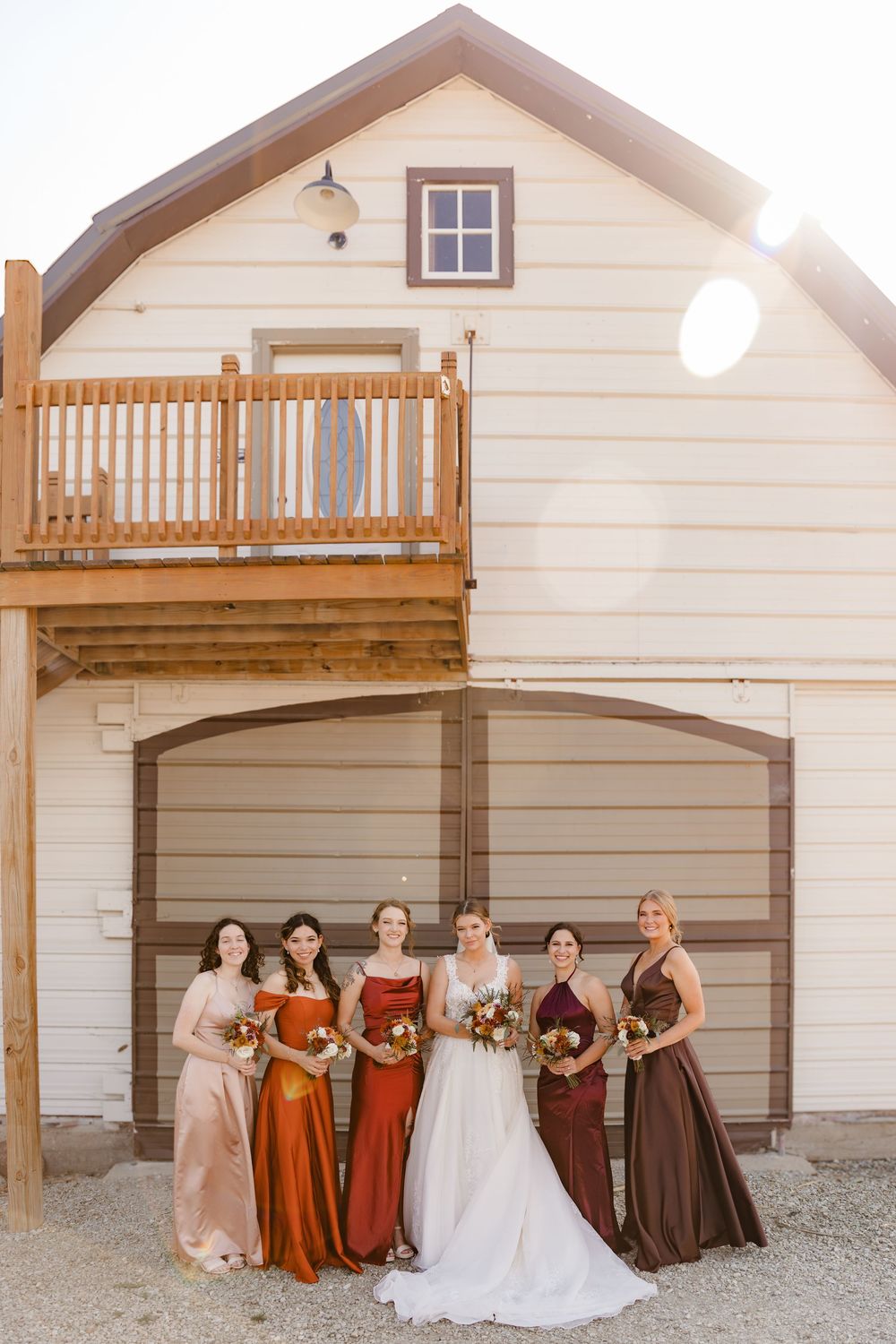 A bridal party wearing fall-toned dresses poses in front of a white barn with a wooden balcony at sunset.