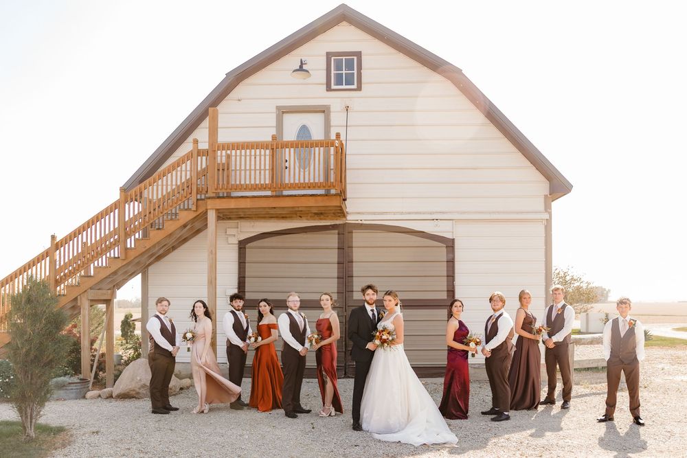 A wedding party poses in front of a white barn at sunset with warm golden lighting creating a romantic atmosphere.