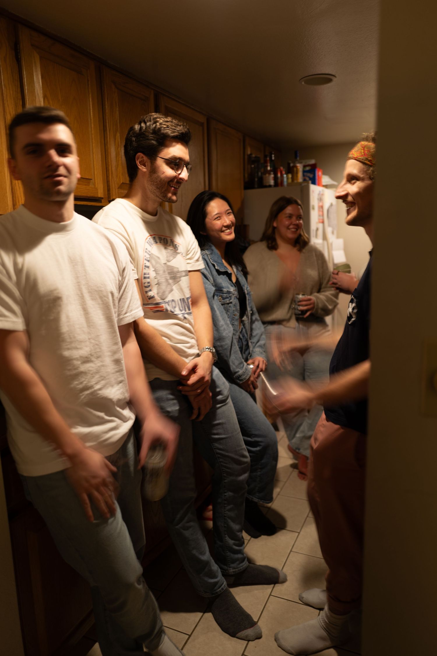Group of five smiling friends gathered together in a warmly lit kitchen at a casual house party.
