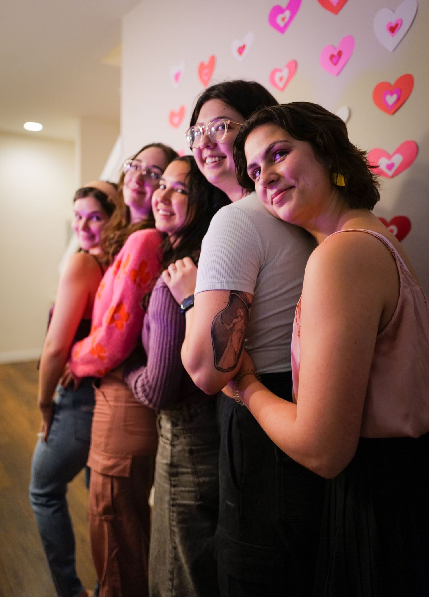 Five women posing together in front of a decorative pink and red heart wall display.