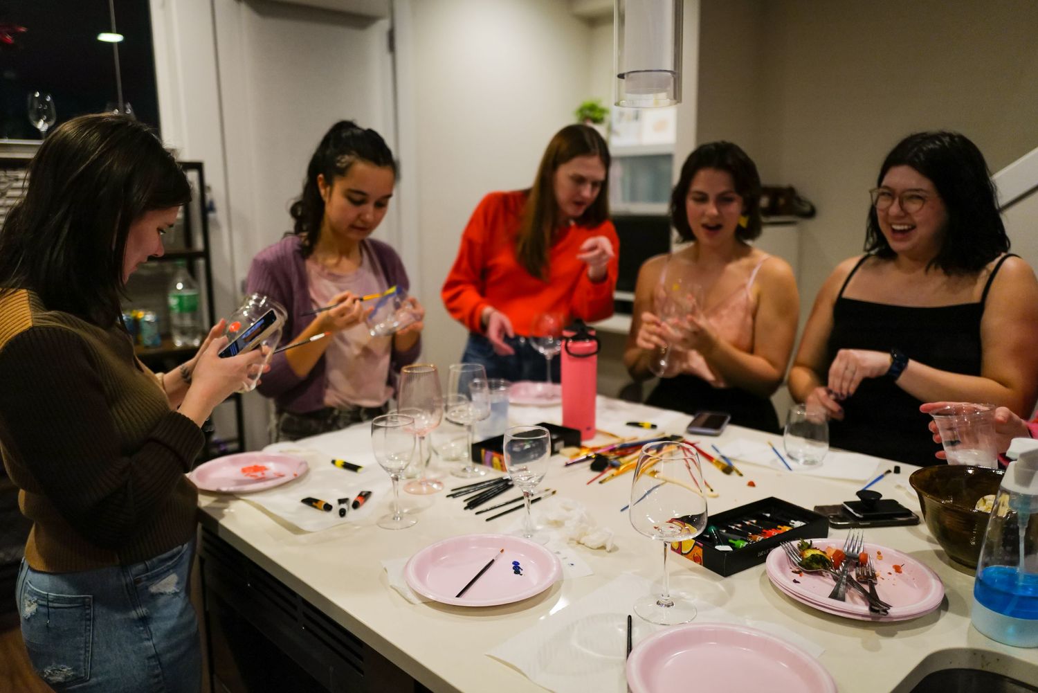 Group of women laughing and painting wine glasses together around a kitchen island at an indoor gathering.