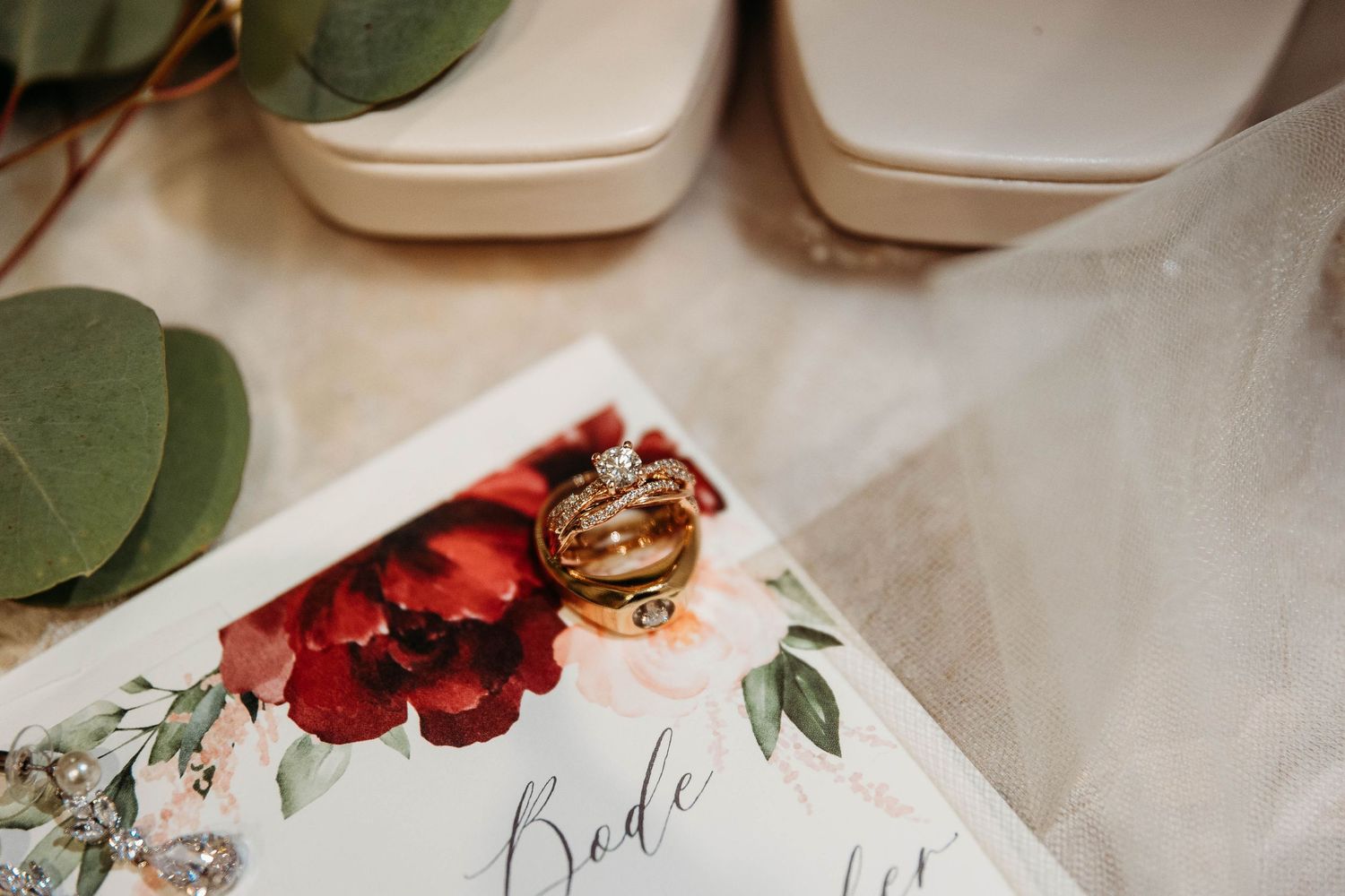 Wedding rings and red floral invitation card with bride text lay beside white shoes on a light surface.