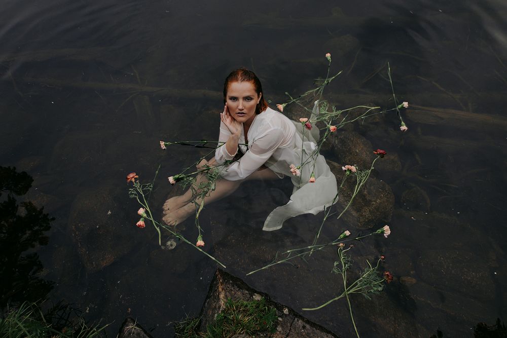 Figure in white clothing submerged in dark water with scattered flowers floating nearby.