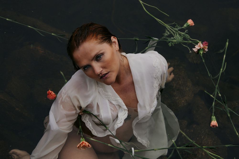 A person in a white button-up shirt poses dramatically with flowers against a dark background.