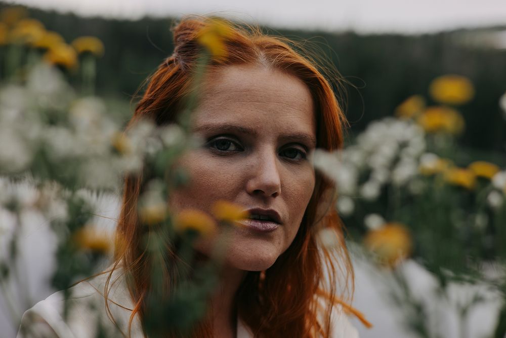 Person with red hair poses among yellow wildflowers in a moody outdoor setting during golden hour.