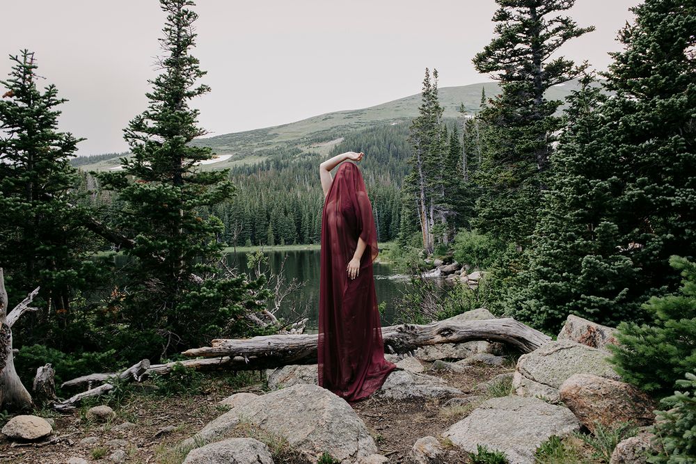 A hiker stands on a rocky mountain overlook surrounded by evergreen trees and distant mountain peaks in the wilderness.