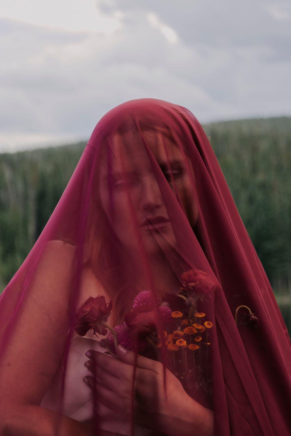 A figure in a deep red veil holds flowers against a blurred natural background.