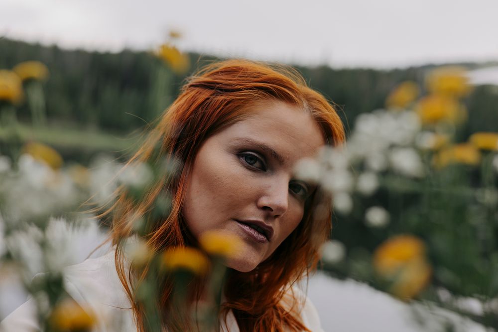 A woman with red hair poses among yellow flowers in a garden setting.