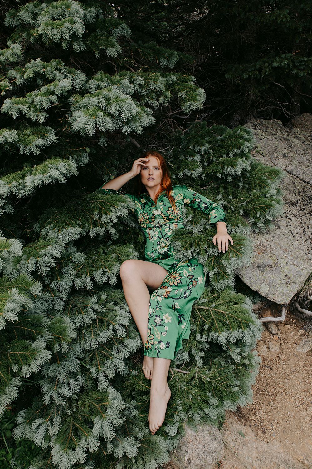 Someone in a green floral dress poses playfully among lush foliage and rocks in a natural outdoor setting.