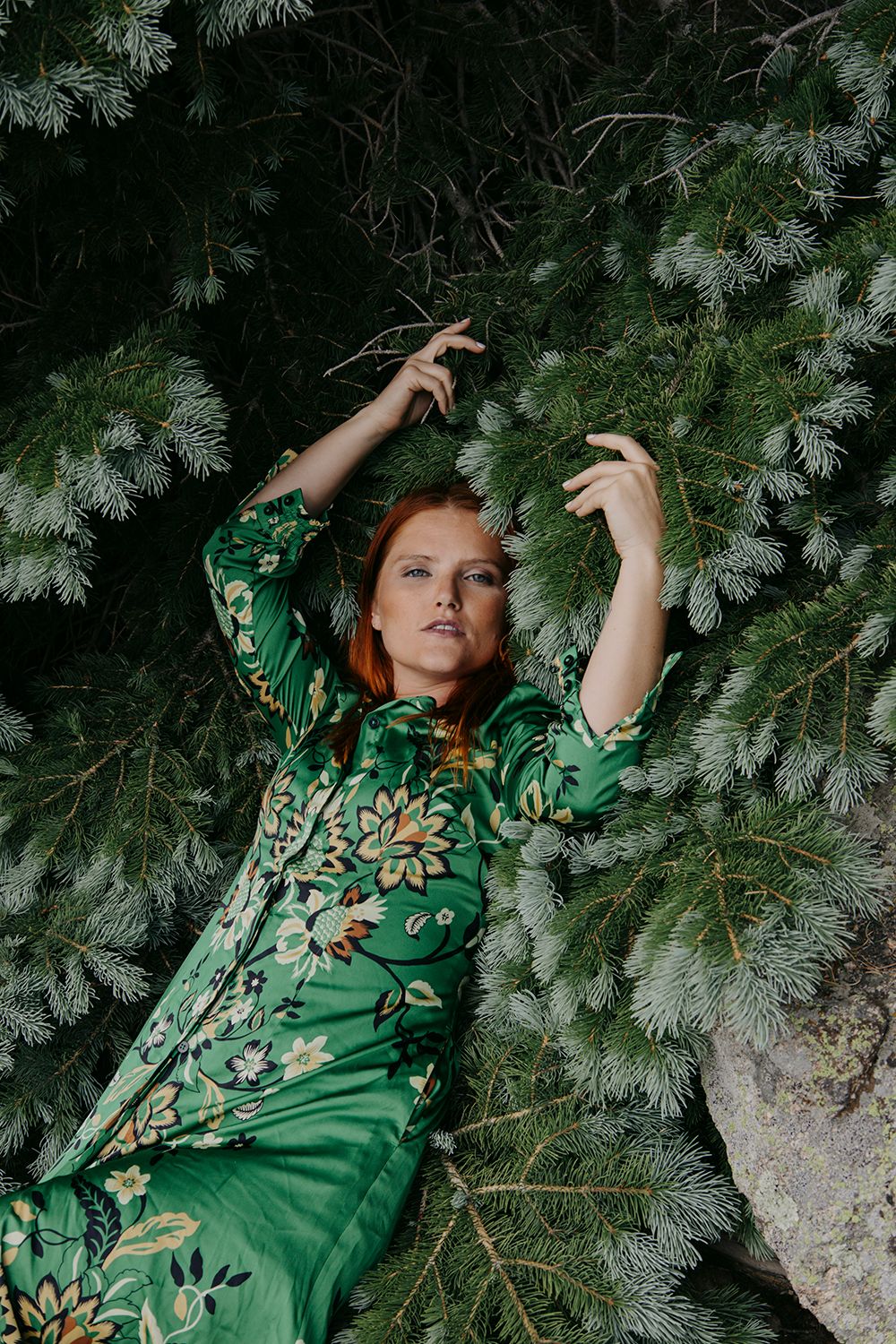 A person in an emerald green floral dress poses artistically among snowy evergreen branches in a moody nature setting.