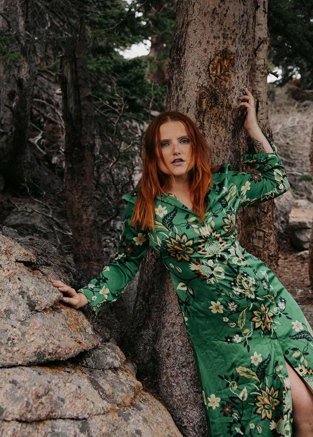 A figure in a flowing emerald green dress poses artistically against tree trunks in a moody forest setting.
