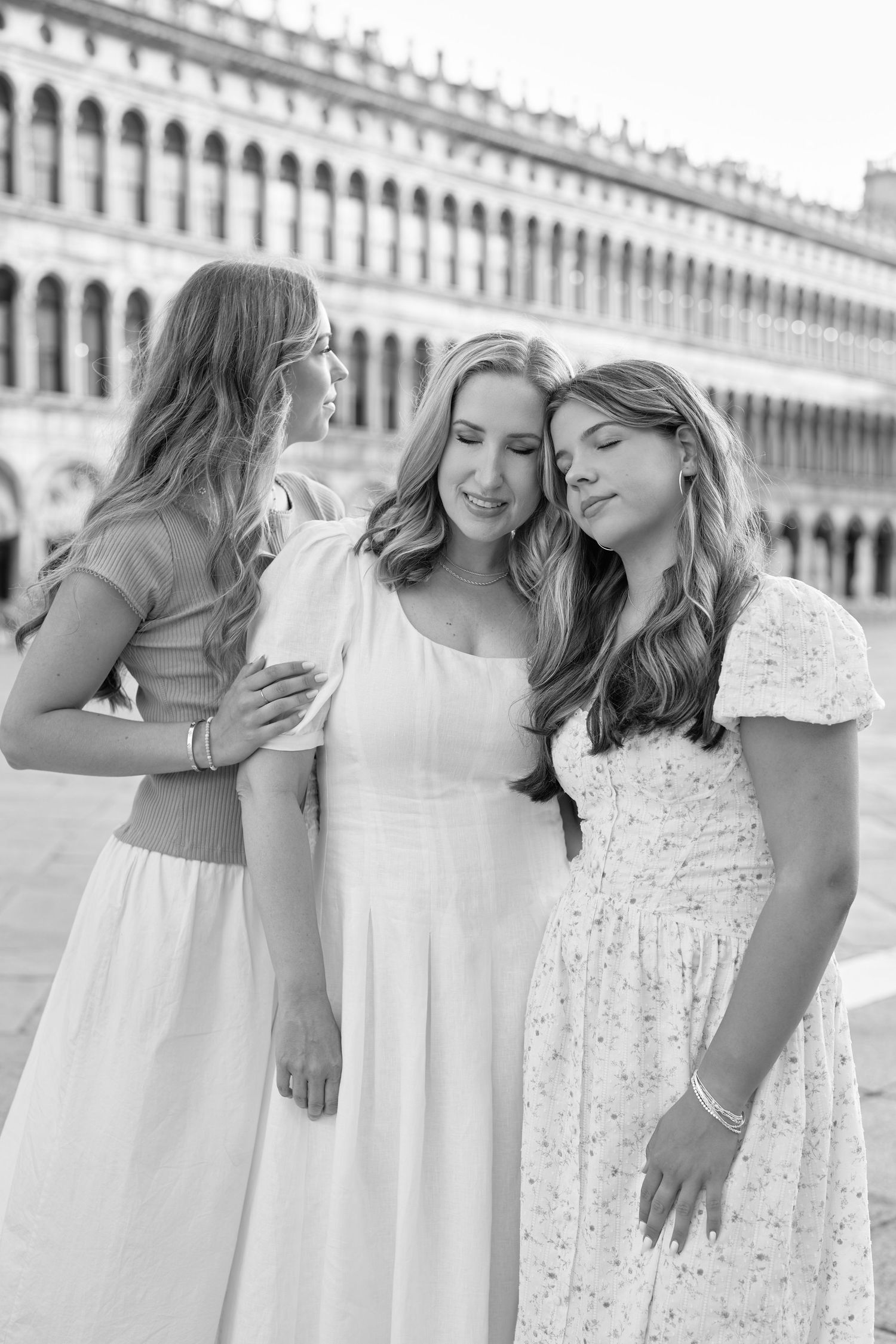 Three women in white dresses laugh and embrace while posing together against an ornate European building facade.