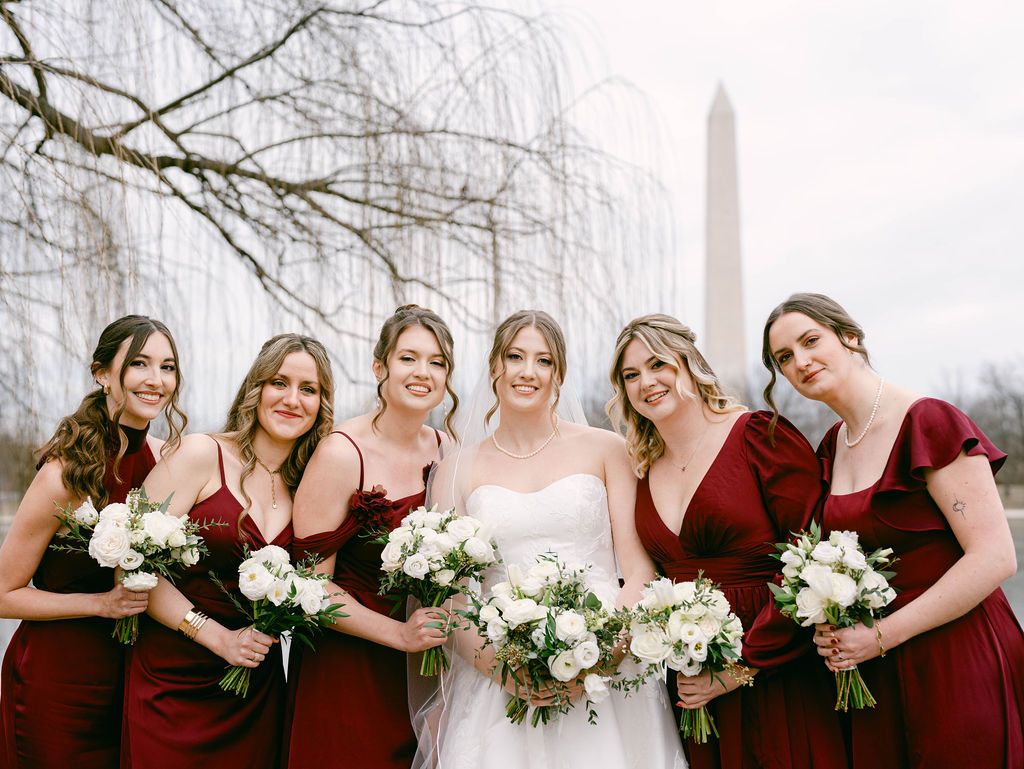Bridesmaids in burgundy dresses pose together holding white floral bouquets on a winter day in front of the Washington Monument in DC.