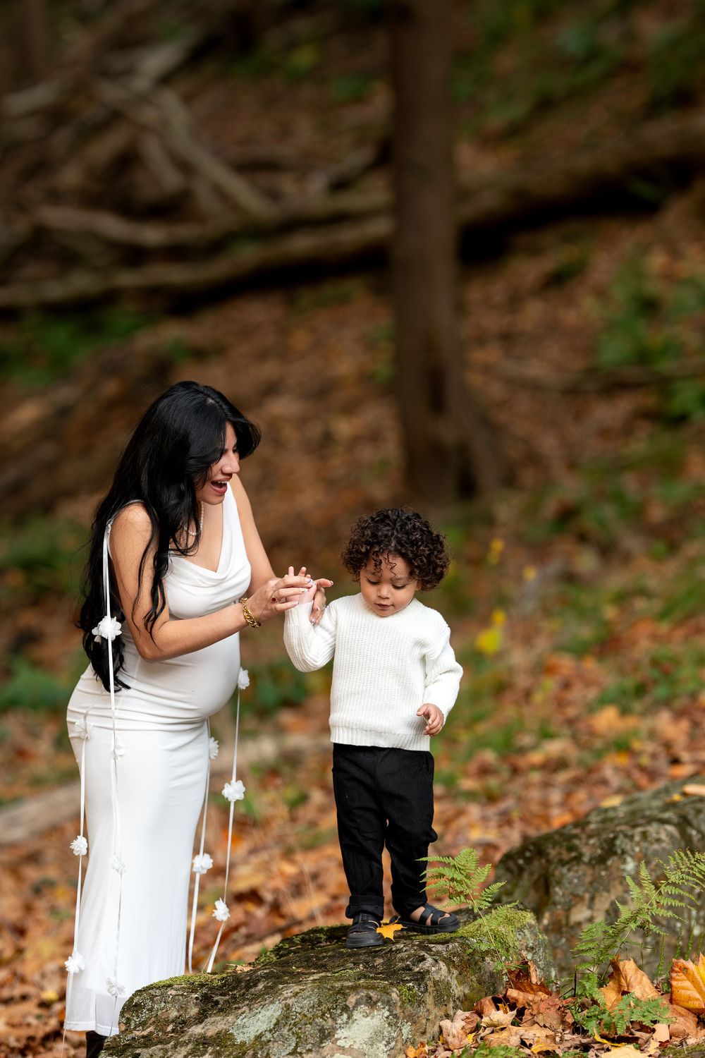 Pregnant mother in white dress holding toddler's hand on rock in fall woods.