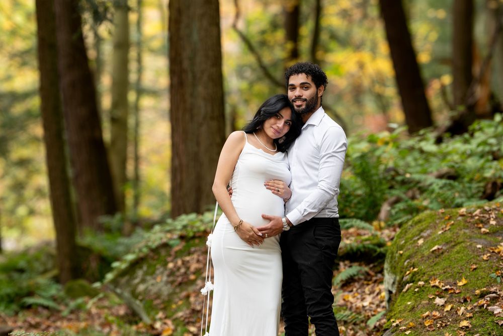 Pregnant woman in white dress embraced by partner in autumn forest maternity portrait.