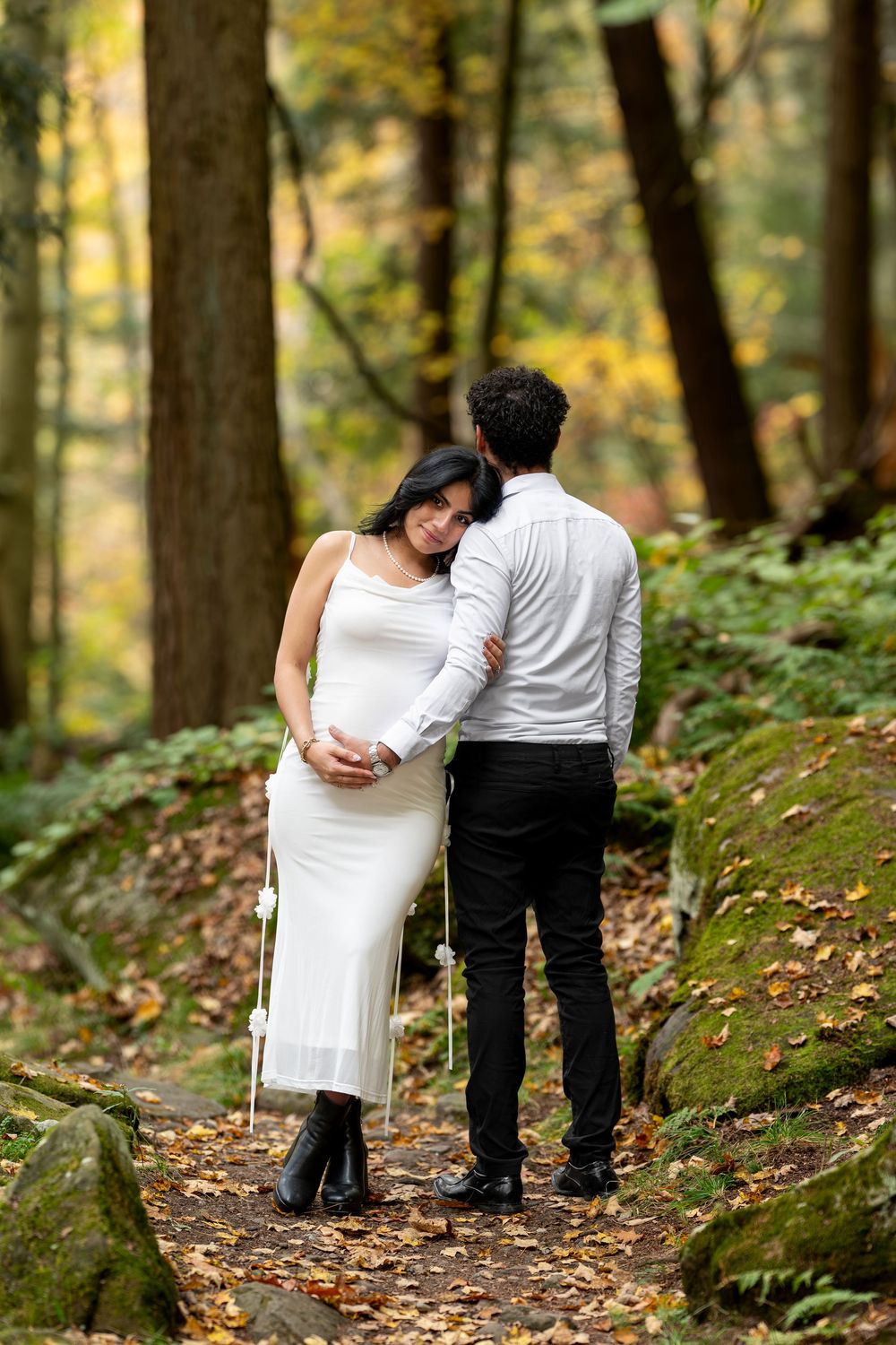 Expecting couple embraces from behind on a wooded trail surrounded by fall foliage.
