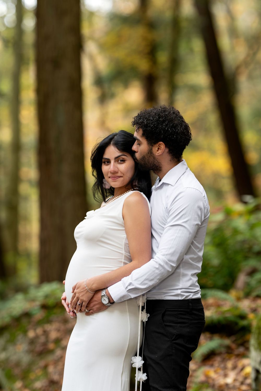 Man tenderly embraces pregnant woman in white dress from behind in a lush autumn forest.