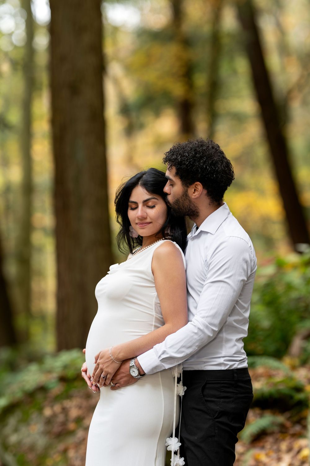 Couple sharing tender moment in autumn forest, man embracing pregnant woman in white dress from behind.