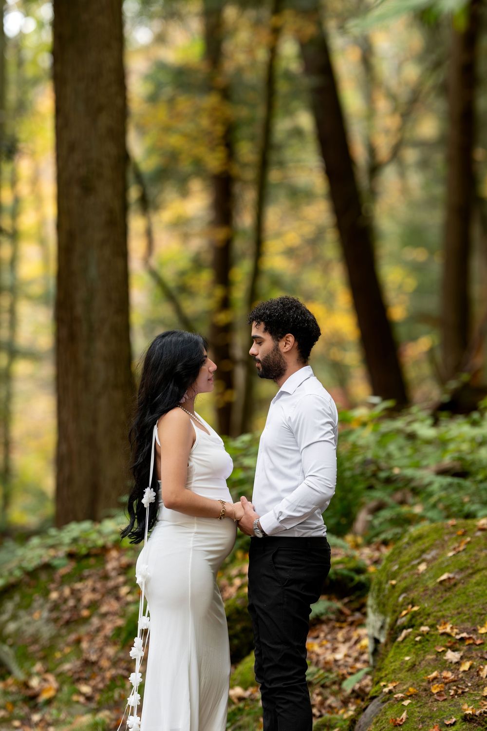 Expecting couple holding hands and gazing at each other on a mossy forest path in fall.