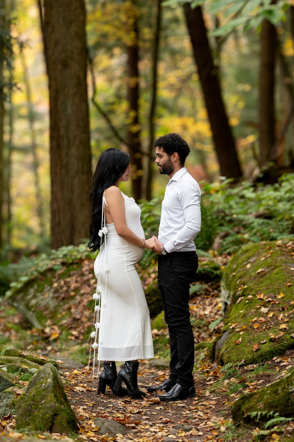 Pregnant woman in white dress and man in white shirt hold hands facing each other in autumn woods.