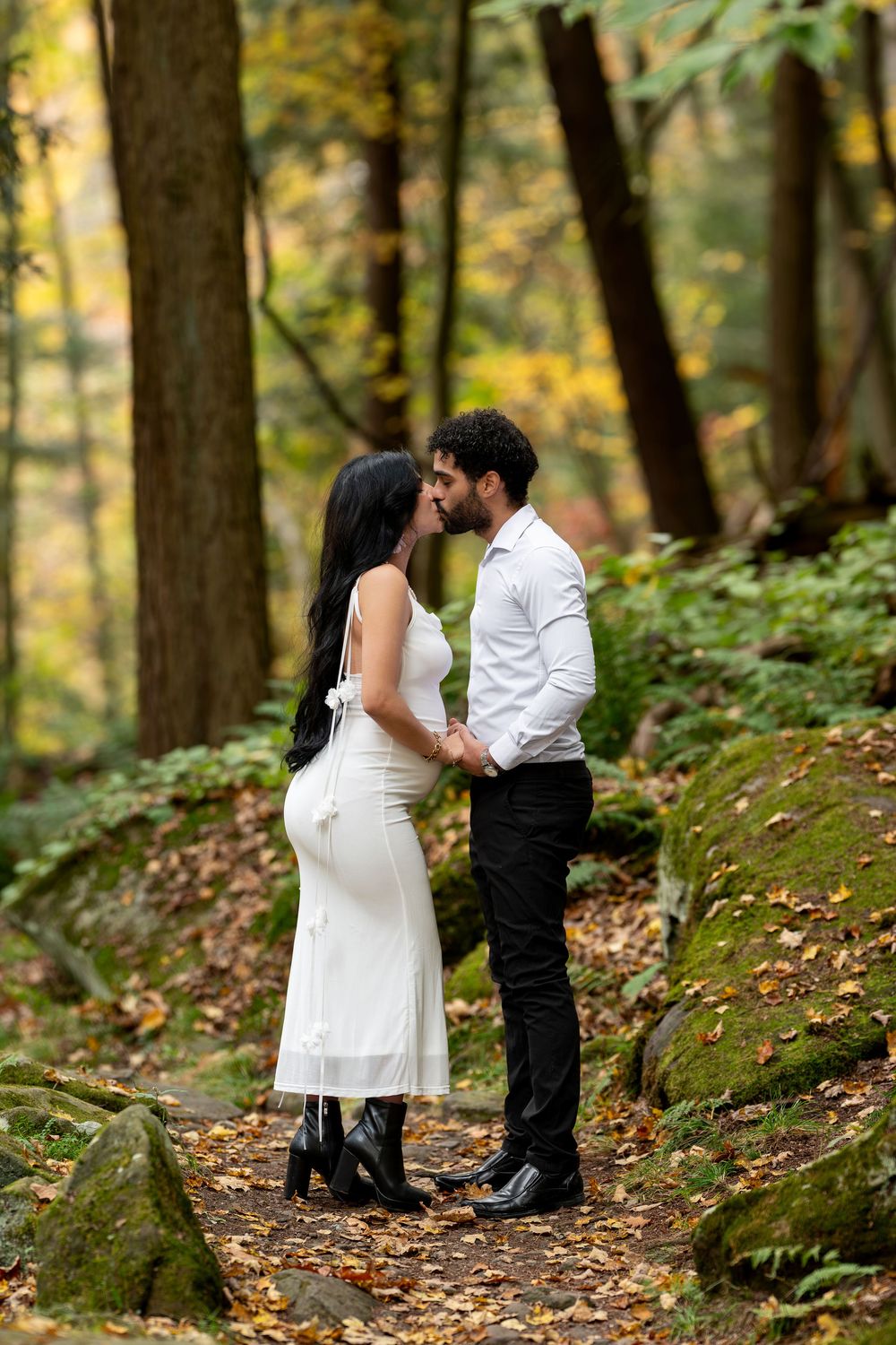 Couple sharing a kiss on a leaf-covered forest trail surrounded by fall foliage.