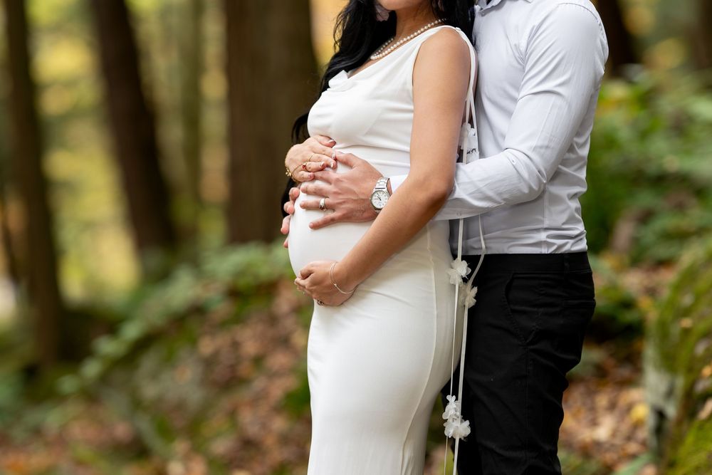 Close-up of pregnant belly with couple embracing outdoors in a lush green forest setting.