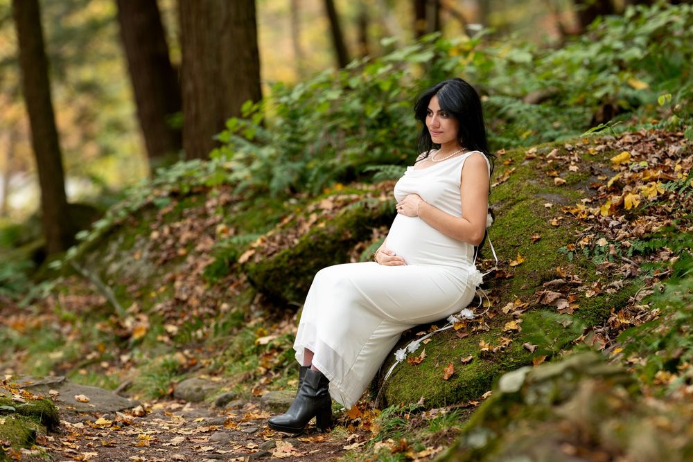 Pregnant woman in white dress relaxing on a bench surrounded by fall leaves in the woods.
