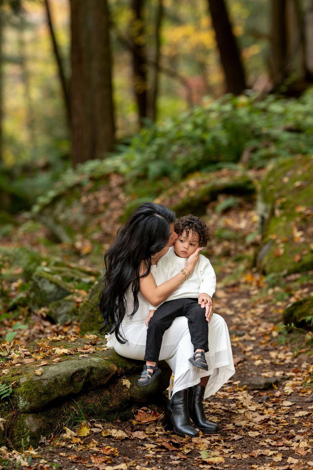 Dark-haired mother kissing toddler's cheek while seated on mossy rocks in autumn woods.