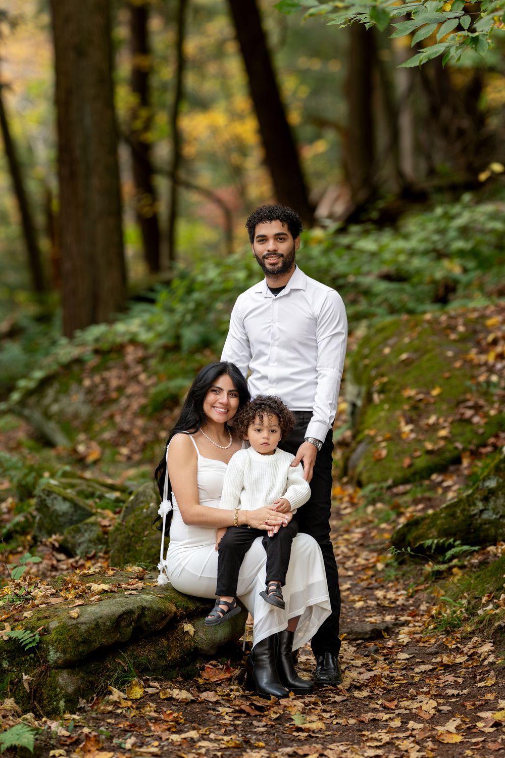Family of three posing on forest rocks surrounded by fall foliage during maternity shoot.