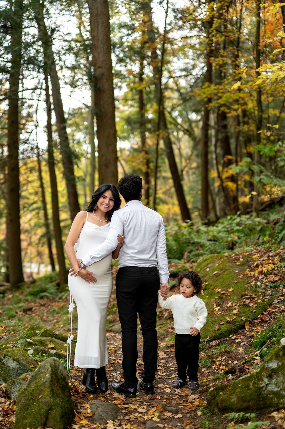 Pregnant woman looks back smiling as family walks along leaf-covered forest trail in fall.