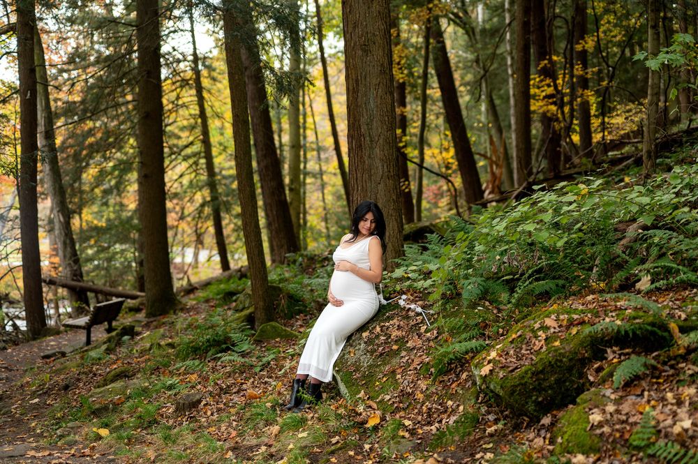 Expecting mother in white outfit posing gracefully on a moss-covered hillside in a lush fall forest.