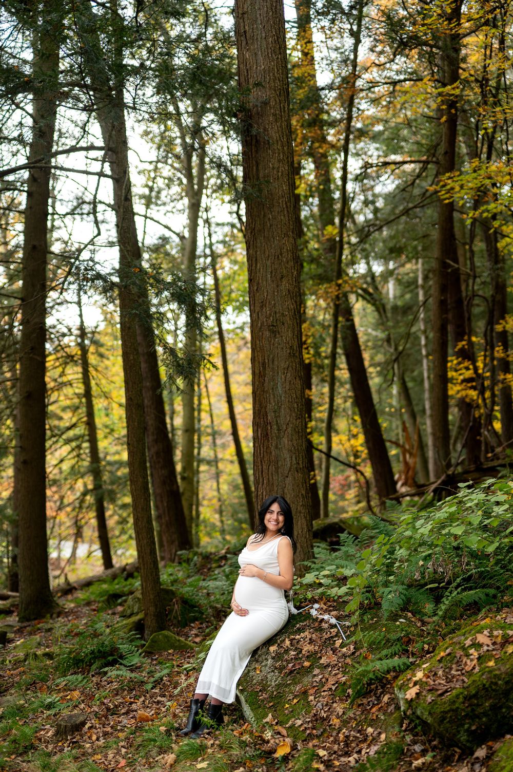 Smiling pregnant woman in white dress cradling her baby bump while leaning against a tall tree in autumn woods.
