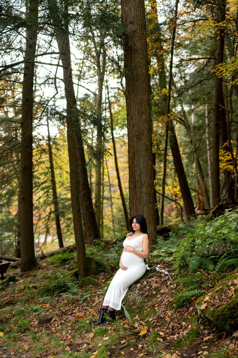 Pregnant woman in white dress leaning against tree in autumn forest.