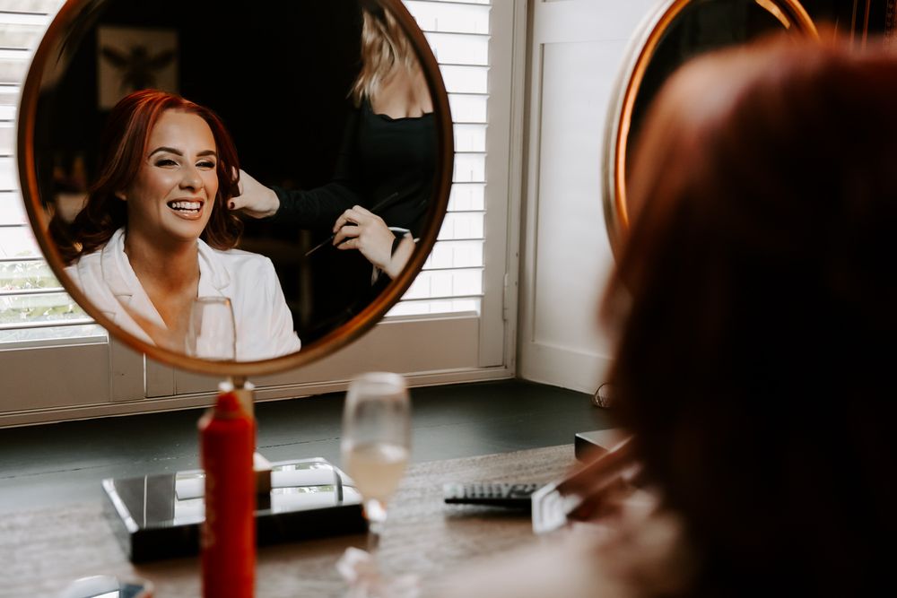 A woman in a black dress twirls gracefully in front of a round copper mirror in a bright sunlit room.